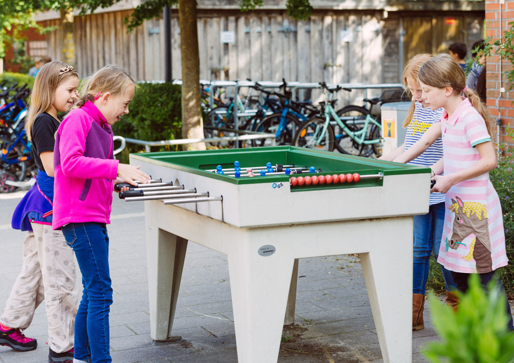 Das Bild zeigt einen Tischfußball auf dem Schulhof der Schule Lehmkuhle. 4 Mädchen haben gerade ein Tisch-Fußball-Match.