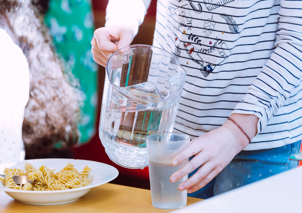 Das Bild zeigt einen Teller mit Nudeln und ein Mädchen, dass ich gerade aus einer Karaffe Wasser in ein Glas gießt. Hier ist gerade Mittagspause in der Mensa des neuen Schulgebäudes.