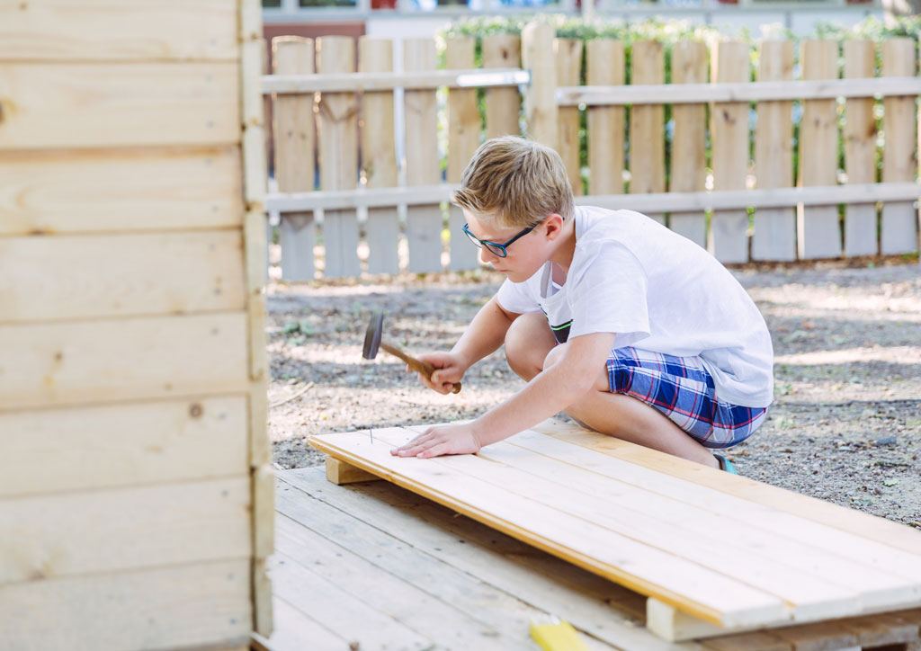 Das Bild zeigt einen Jungen, der mit einem Hammer einen Nagel in ein Brett nagelt. Die Kinder haben in der Ganztagsbetreuung die Möglichkeit viel mit Holz zu bauen.