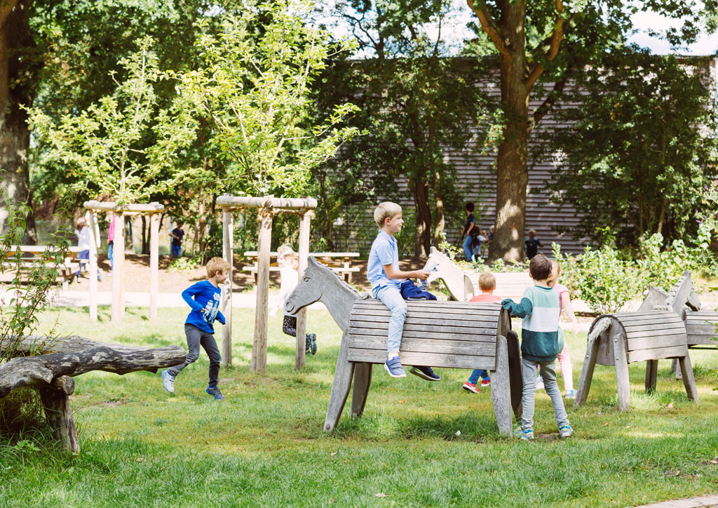 Das Bild zeigt einen Teil des Spielbereichs draußen auf dem Schulhof der Schule Lehmkuhle. Davor befinden sich mehrere große Holzpferde und spielende Kinder.