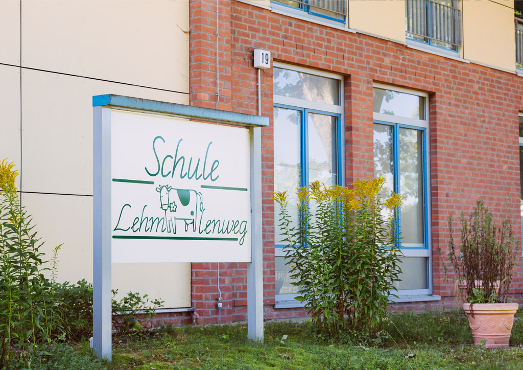 Das Bild zeigt das große Eingangsschild der Schule Lehmkuhle mit dem Kuh-Logo. In dem hinteren Gebäude befindet sich die Aula.