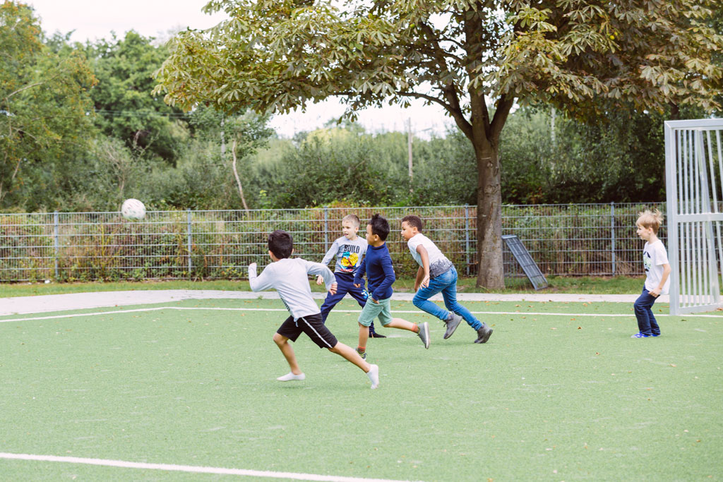 Das Bild zeigt einen Teil des Fußballplatzes der Schule Lehmkuhle. Dort spielen fünf Jungs Fußball.