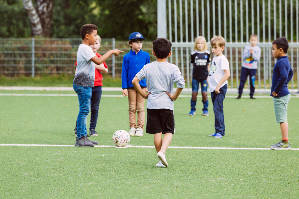 Das Bild zeigt einen Teil des Fußballplatzes der Schule Lehmkuhle. Dort spielen einige Jungs Fußball und es wird gerade diskutiert, wer den Einwurf macht.