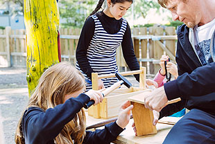 Das Bild zeigt zwei Kinder mit einem Betreuuer aus der GBS. Die Kinder basteln unter Anleitung etwas aus Holz.